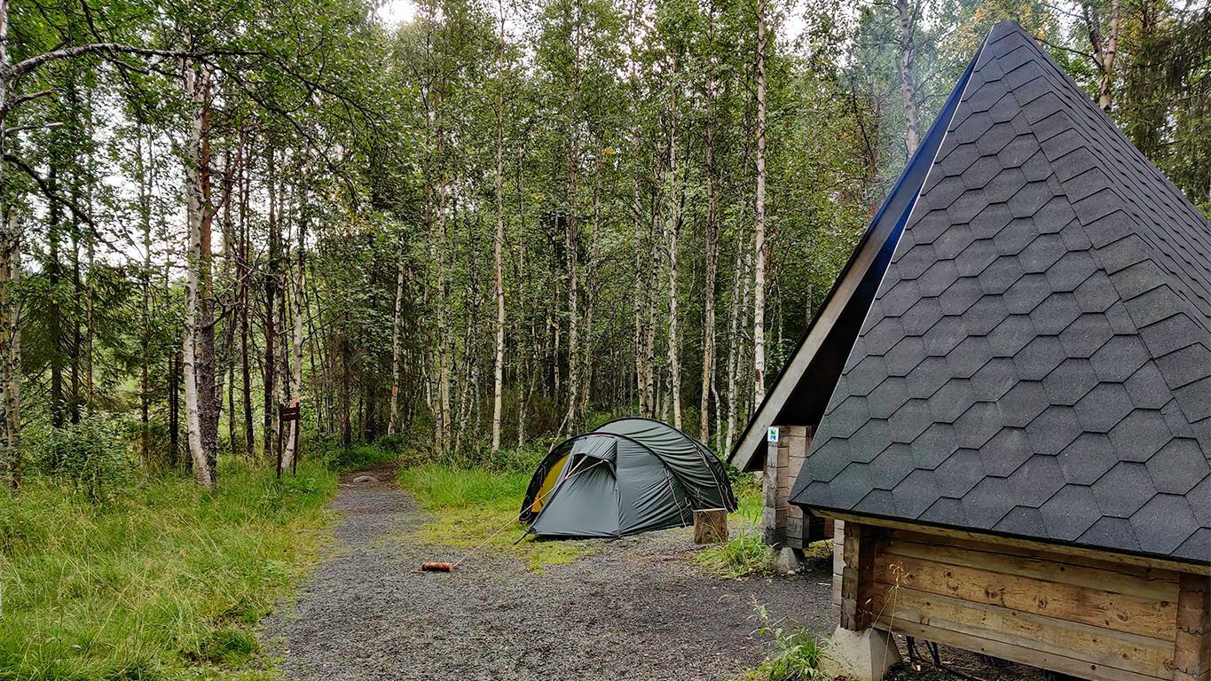 My 4-person green tent next to a large wooden shelter, from where smoke tufts out from an open fire. The camp has gravel ground srrounded by long grass and 10+ meter tall birch trees.