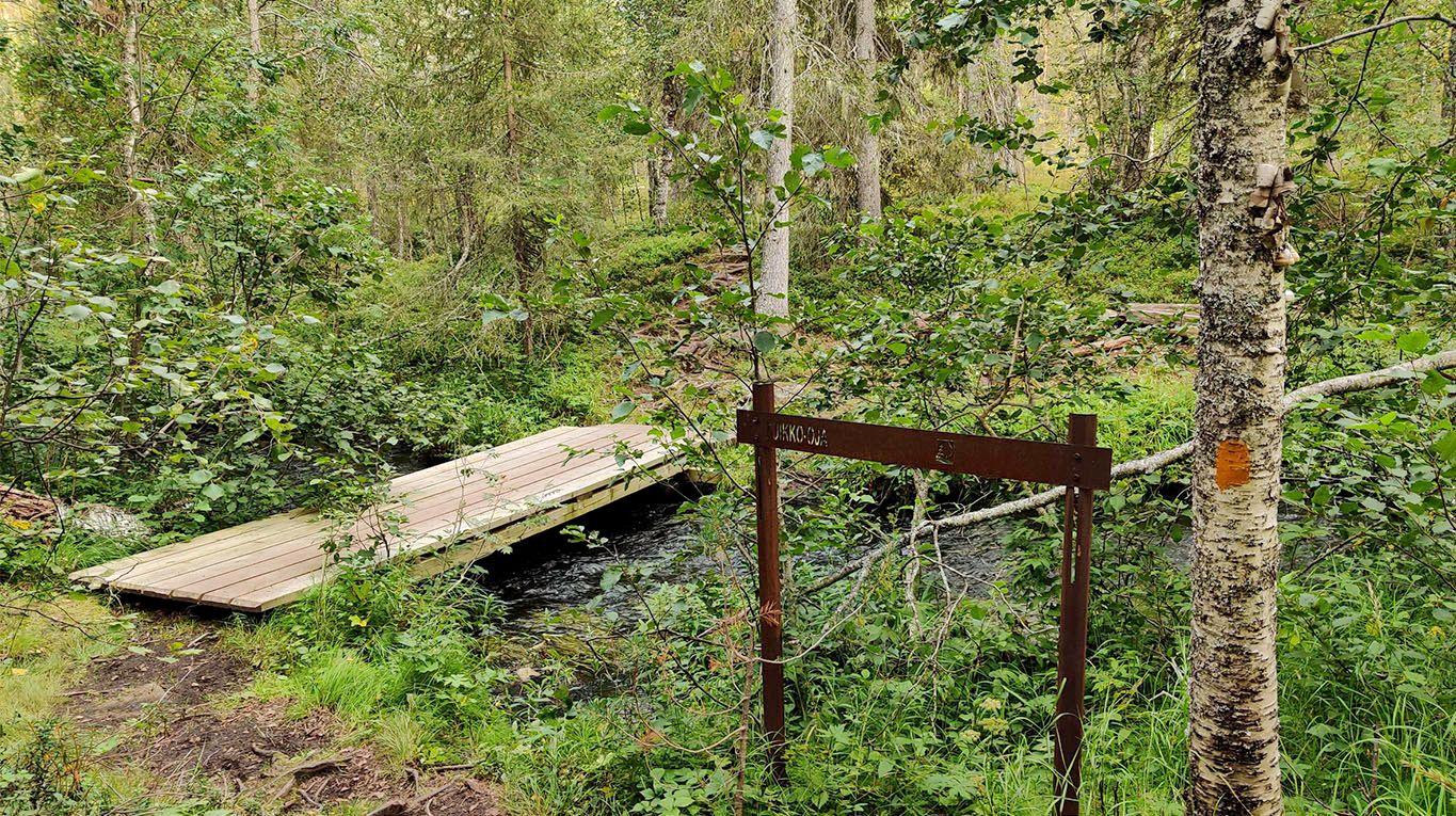 A sturdy plank bridge across a wide stream with trees surrounding the stream and a signpost indicating the name.