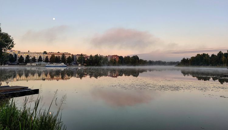 Reflections of buildings, clouds, on mirror-like lake surface, with mist rolling slowly.