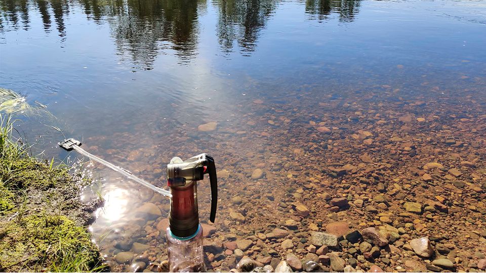 A heavy handheld water purifier pumping water from a clear river on a sunny August day.
